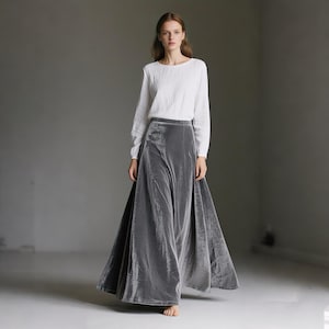 Puede incluir: Una mujer con una falda larga de terciopelo gris y una blusa blanca. La falda es de cintura alta y tiene una silueta amplia y fluida. El conjunto es sencillo y elegante, adecuado para diversas ocasiones. La falda mide aproximadamente 100 cm de largo.
