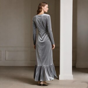 May include: A long, silver-gray velvet dress with long sleeves and a ruffled hem. The dress has a V-neck and a fitted bodice that flows into a flared skirt. The model is wearing gold-colored heels.