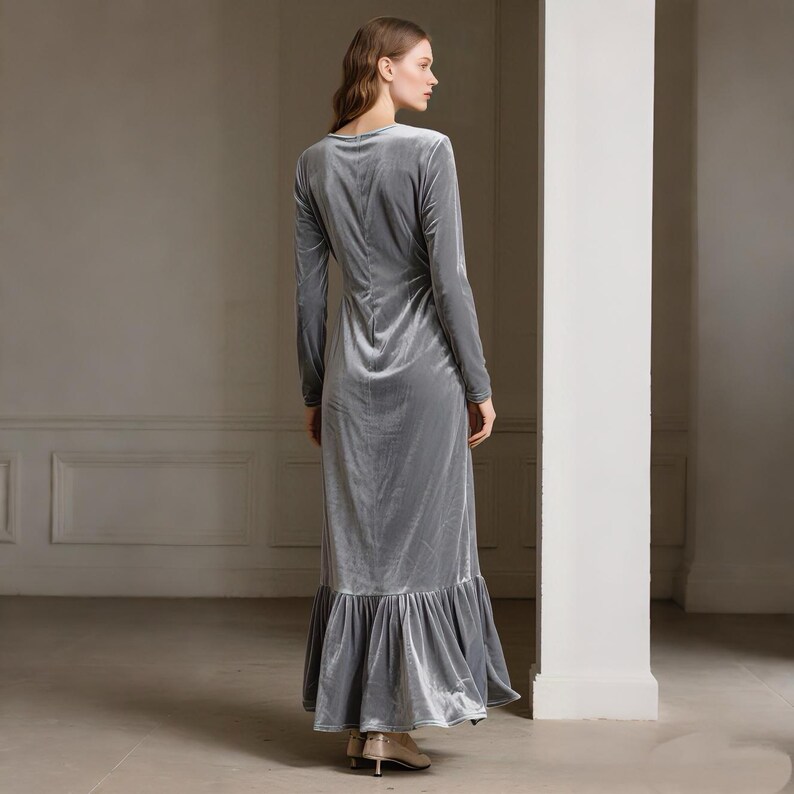 May include: A long, silver-gray velvet dress with long sleeves and a ruffled hem. The dress has a V-neck and a fitted bodice that flows into a flared skirt. The model is wearing gold-colored heels.