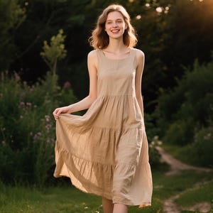 Könnte beinhalten: Ein ärmelloses, beigefarbenes Leinenkleid mit einem gestuften Rock. Das Kleid hat einen runden Ausschnitt und reicht bis zur Mitte der Wade. Die Frau geht in einem grasbewachsenen Bereich mit Bäumen und Sonnenlicht im Hintergrund.