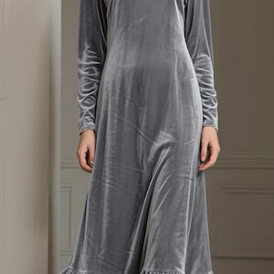 May include: A long, flowing, gray velvet dress with long sleeves and a round neckline. The dress features a ruffled hemline and is floor-length. The fabric has a soft sheen, and the dress appears to be well-tailored.