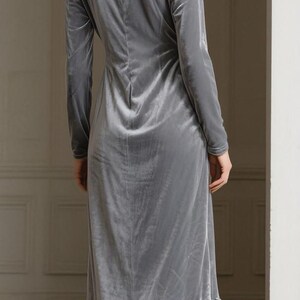 May include: A long, light gray velvet dress with long sleeves and a ruffled hem. The dress has a V-neck and a fitted silhouette. The model is wearing the dress, seen from the back.