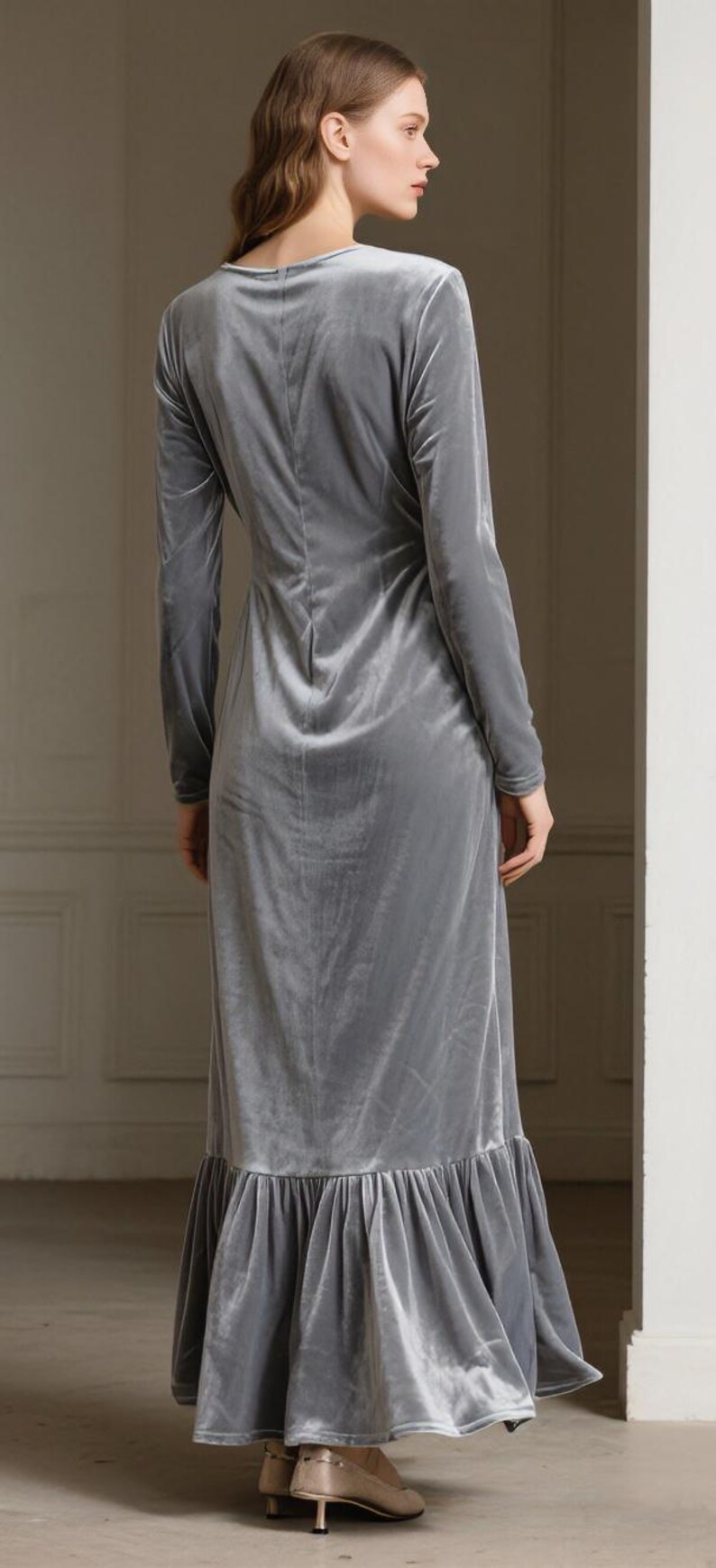 May include: A long, light gray velvet dress with long sleeves and a ruffled hem. The dress has a V-neck and a fitted silhouette. The model is wearing the dress, seen from the back.