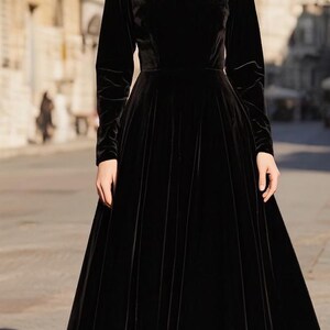 Puede incluir: Un vestido largo de terciopelo negro con mangas largas y cuello redondo. El vestido tiene un corpi&ntilde;o ajustado y una falda fluida. La modelo est&aacute; de pie en una calle adoquinada frente a un edificio.