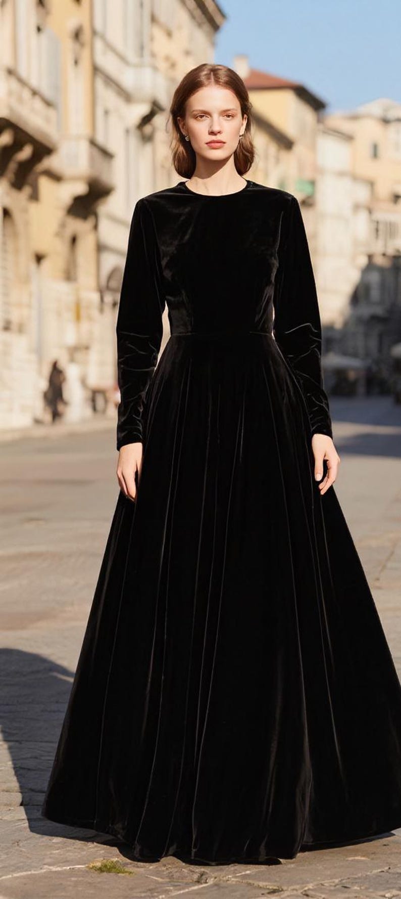 Puede incluir: Un vestido largo de terciopelo negro con mangas largas y cuello redondo. El vestido tiene un corpi&ntilde;o ajustado y una falda fluida. La modelo est&aacute; de pie en una calle adoquinada frente a un edificio.
