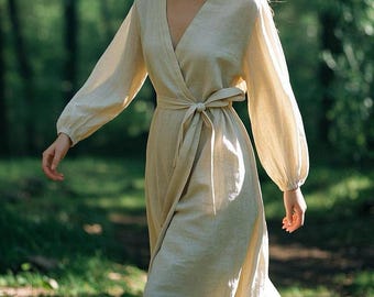 May include: A beige wrap dress featuring long, loose sleeves with gathered cuffs, a deep V-neckline, and a matching fabric tie belt at the waist. The dress has a floor-length skirt and is constructed from a solid, light-colored woven fabric.
