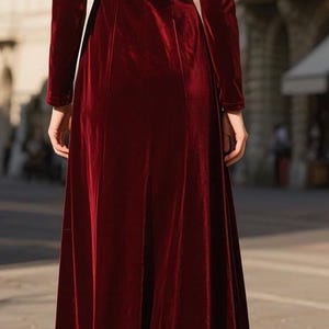 May include: A long, burgundy velvet dress with long sleeves. The dress has a high neckline and a flowing skirt. The model is standing on a cobblestone street in front of a building.