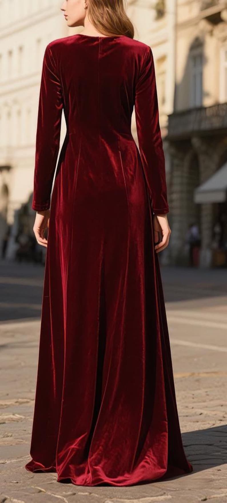 May include: A long, burgundy velvet dress with long sleeves. The dress has a high neckline and a flowing skirt. The model is standing on a cobblestone street in front of a building.