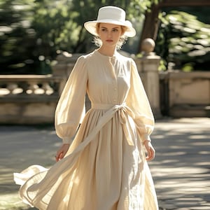 Puede incluir: Una mujer con un vestido beige de manga larga con un cinturón blanco y un sombrero blanco. El vestido tiene un escote redondo y una falda fluida. La mujer está de pie en un entorno de parque.