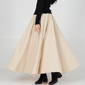 May include: A beige, full-length skirt with a gathered waistline. The skirt is flowing and has a wide, circular shape.