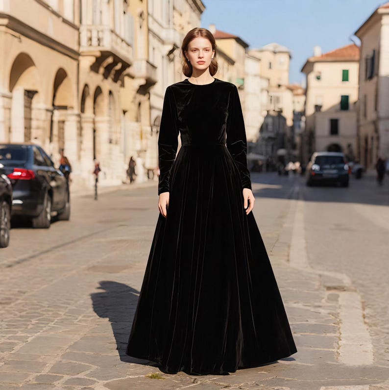 Puede incluir: Una mujer con un vestido largo de terciopelo negro con mangas largas y cuello redondo. El vestido tiene una falda amplia. El escenario es una calle europea con edificios y un coche.