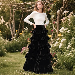 May include: A woman wearing a white long-sleeve top and a black tiered velvet skirt. The skirt has multiple layers of ruffles. The background features a garden with flowers and trees.