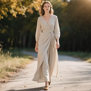 May include: A long, beige wrap dress with a V-neck and long sleeves. The dress is made of a lightweight fabric and has a knotted detail at the waist. The model is walking on a dirt path.