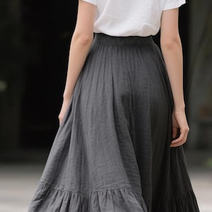Pode incluir: Uma t-shirt branca de manga curta combinada com uma saia maxi comprida em cinzento escuro. A saia apresenta um design franzido de cintura subida com uma costura horizontal na bainha, criando uma silhueta larga e fluida at&eacute; ao ch&atilde;o.