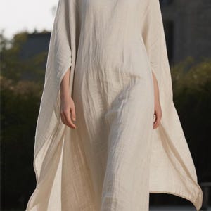 May include: A flowing, off-white linen dress with a round neckline and wide sleeves. The dress is floor-length and appears lightweight, suitable for warm weather. The model is walking on a light-colored surface.