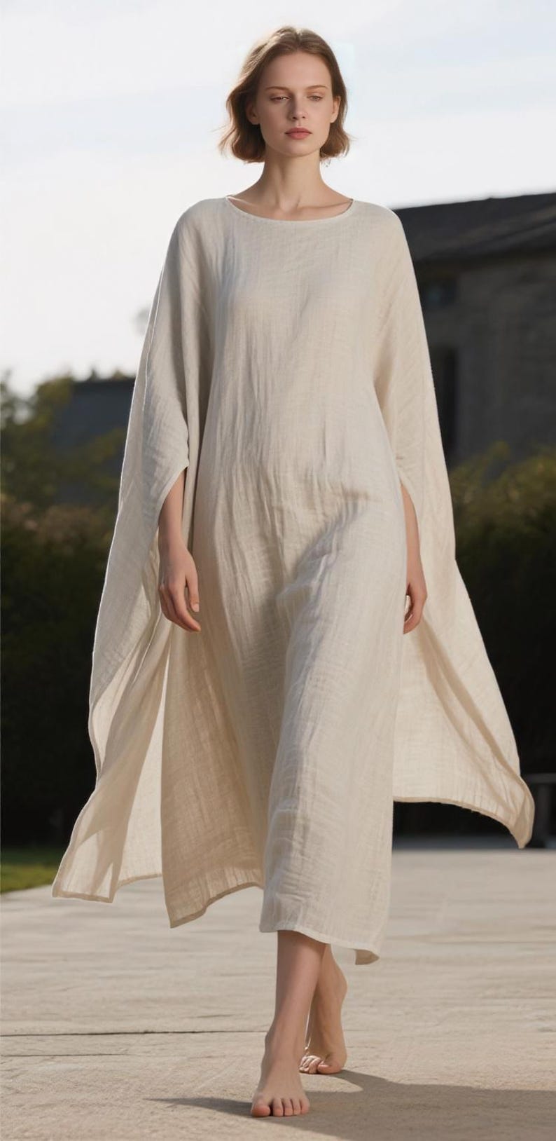 May include: A flowing, off-white linen dress with a round neckline and wide sleeves. The dress is floor-length and appears lightweight, suitable for warm weather. The model is walking on a light-colored surface.