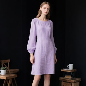 May include: A knee-length lavender dress with long, billowy sleeves and a round neckline. The garment features a fitted bodice that transitions into a slightly flared skirt, crafted from a textured fabric with a subtle weave pattern throughout.