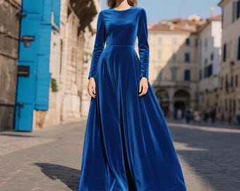 May include: A long, flowing, royal blue velvet dress with long sleeves. The dress has a fitted bodice and a full skirt that flares out from the waist. The model is standing on a cobblestone street.