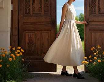 May include: A woman in a white linen dress stands in front of a wooden door with carved details. She is holding the door open with her right hand. There are yellow flowers in the foreground.