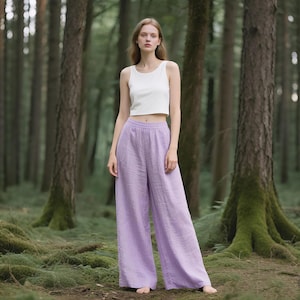 May include: A woman wearing a white tank top and wide-leg lavender pants stands in a forest. The pants are a light purple color and appear to be made of linen or a similar lightweight fabric. The background features tall trees and green foliage.