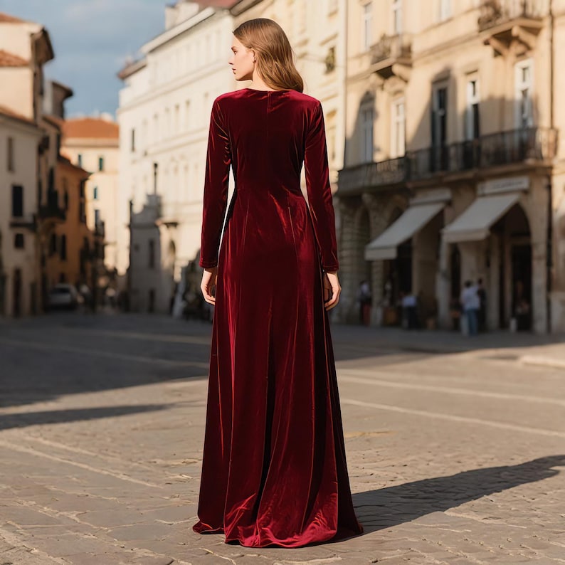 May include: A long, burgundy velvet gown with long sleeves and a flowing skirt. The dress has a simple, elegant design and is worn by a person standing on a cobblestone street.