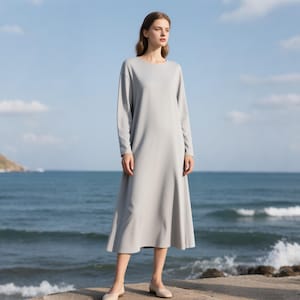 May include: A light gray long-sleeve midi dress with a round neckline and a relaxed, flowing silhouette that falls below the knees. The garment features a smooth, solid-colored fabric with long sleeves and a simple, unadorned design throughout.
