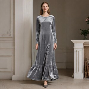 May include: A long, gray velvet dress with long sleeves and a round neckline. The dress has a flowing skirt with a ruffled hem. The model is wearing gray heels. The dress is suitable for formal occasions.