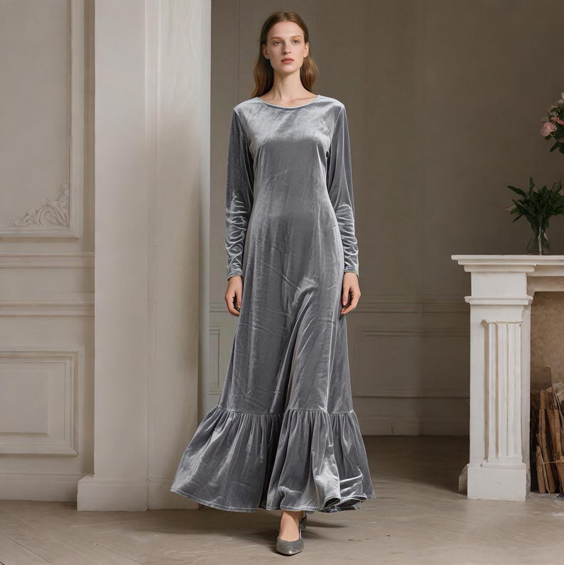 May include: A long, gray velvet dress with long sleeves and a round neckline. The dress has a flowing skirt with a ruffled hem. The model is wearing gray heels. The dress is suitable for formal occasions.