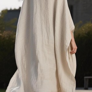 May include: A long, flowing, cream-colored linen garment with wide sleeves. The dress has a simple, elegant design and drapes loosely. The model is wearing the dress outdoors, with a blurred background of greenery and a building.