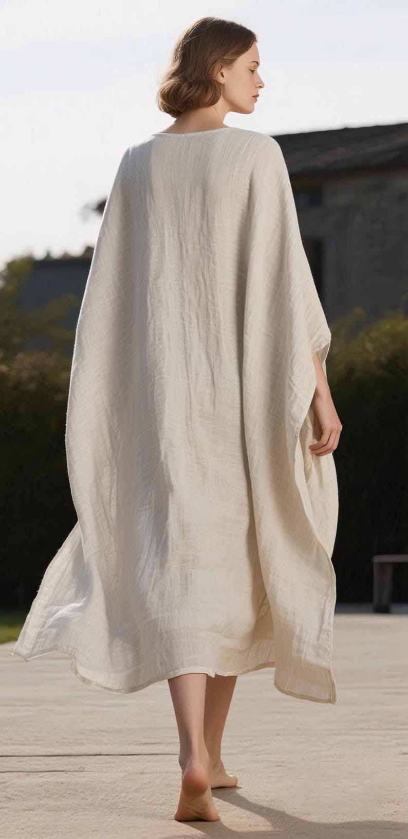May include: A long, flowing, cream-colored linen garment with wide sleeves. The dress has a simple, elegant design and drapes loosely. The model is wearing the dress outdoors, with a blurred background of greenery and a building.