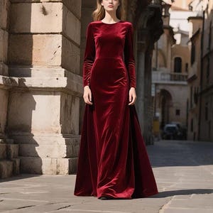 May include: A long, burgundy velvet gown with long sleeves and a flowing skirt. The dress has a simple, elegant design and is worn by a person standing on a cobblestone street. The background features stone architecture and a glimpse of a city street.