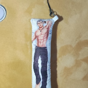 Peut inclure: Un oreiller blanc avec une image imprimée d'un homme musclé portant un pantalon noir. L'oreiller est muni d'une sangle noire.