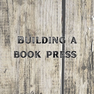 Op de afbeelding: Een houten bord met de tekst "BUILDING A BOOK PRESS" in zwarte letters. Onder de tekst staat een logo met de tekst "Hand Crafted By Dallas Ryan Bennett".