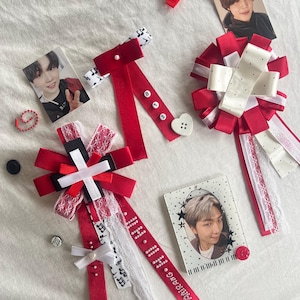 May include: Three decorative ribbon rosettes and bows in red, white, and black, featuring lace trim, heart-shaped buttons, letter beads spelling BTS, and musical note patterns. Included are two small portrait cards of individuals and a red ball chain.
