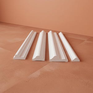 Four white wall molding pieces arranged on a terracotta-colored surface. The molding has decorative detailing and a smooth, clean finish. The pieces are angled, showcasing their design and profile.