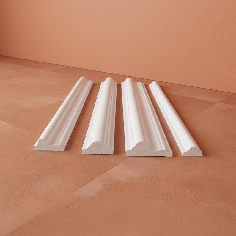 Four white wall molding pieces arranged on a terracotta-colored surface. The molding has decorative detailing and a smooth, clean finish. The pieces are angled, showcasing their design and profile.