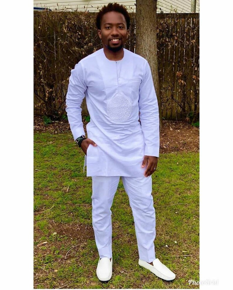 May include: A white, long-sleeved tunic and matching pants set. The tunic features a round neckline and a decorative embroidered design on the front. The outfit is paired with white slip-on shoes. The person is standing on green grass.