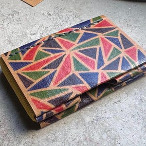 Può includere: Un portafoglio in pelle fatto a mano con un motivo geometrico rosso, blu, verde e rosa. Il portafoglio è piegato e cucito con un filo a contrasto. I bordi sono di colore marrone chiaro.
