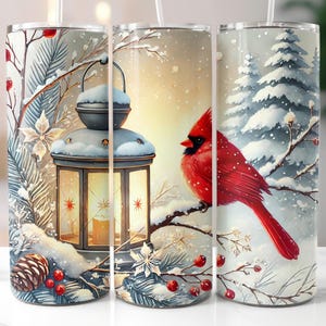 May include: A stainless steel tumbler featuring a winter scene with a red cardinal perched on a snow-covered branch. The design includes a lantern, pine cones, and berries, evoking a cozy, seasonal theme. The tumbler is designed for cold beverages.