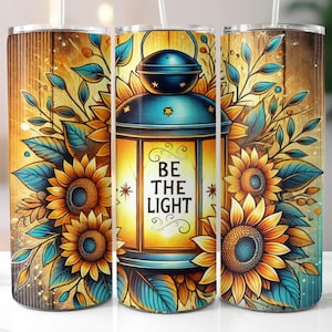 May include: A tumbler with a floral design featuring sunflowers and teal leaves. The center of the tumbler has a teal lantern with the words "BE THE LIGHT" in white script. The background has a wood grain pattern.