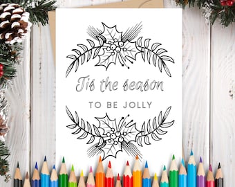 May include: A white card with the text "Tis the season to be jolly" in a decorative font, framed by holly and leaf illustrations. The card is surrounded by colored pencils and holiday greenery.