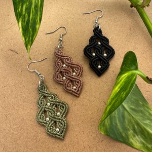 Peut inclure: Trois boucles d'oreilles en macramé de couleur vert olive, vieux rose et noir. Chaque boucle présente un motif de cordons noués en forme de losange avec de petites perles métalliques argentées intégrées, suspendues à des crochets en métal argenté.
