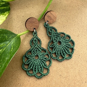 Peut inclure: Une paire de boucles d'oreilles en macramé vert foncé. Chaque boucle d'oreille présente un motif floral suspendu à un petit clou rond en bois. Les boucles d'oreilles sont faites à la main et présentées sur un fond neutre.
