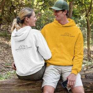 May include: A couple wearing white and yellow hoodies with the text "ADVENTURE JUNKIE" and a mountain graphic. The white hoodie has additional text: "Be brave, wild and free." They are sitting on a log in a forest setting.