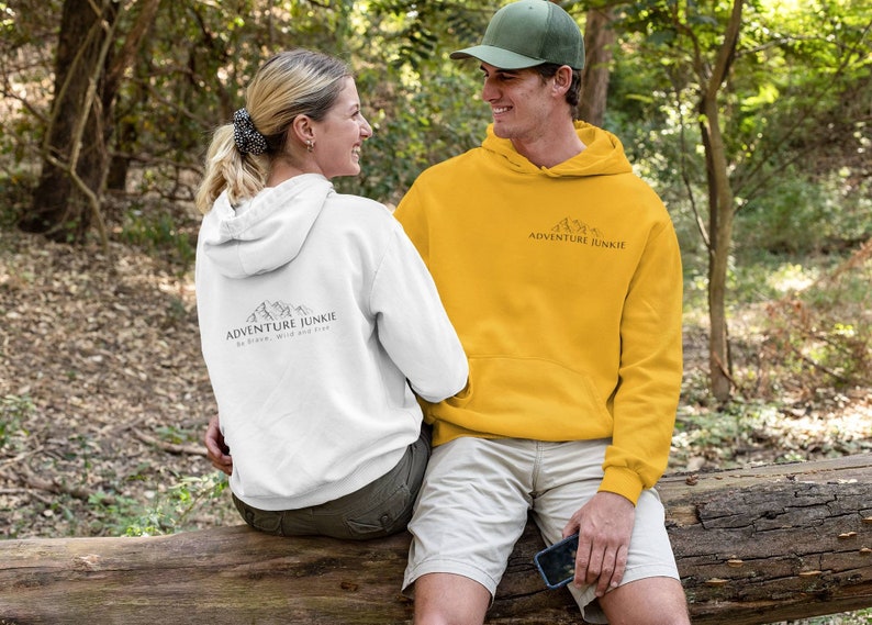 May include: A couple wearing white and yellow hoodies with the text "ADVENTURE JUNKIE" and a mountain graphic. The white hoodie has additional text: "Be brave, wild and free." They are sitting on a log in a forest setting.