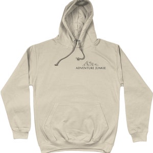 May include: A tan hoodie with a drawstring hood and a front pocket. The hoodie features a small mountain graphic and the text "ADVENTURE JUNKIE" printed in black. The hoodie is made of a soft material.