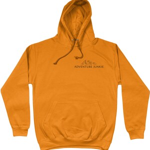 May include: Mustard-colored hoodie with a drawstring hood and a front pocket. The hoodie features a small mountain graphic and the text "ADVENTURE JUNKIE" printed in black. A casual, comfortable garment for outdoor enthusiasts.