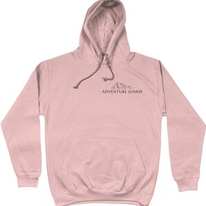 May include: A light pink hoodie with a drawstring hood and a kangaroo pocket. The hoodie features a small mountain graphic and the text "ADVENTURE JUNKIE" printed in black. The hoodie is made of a soft material.