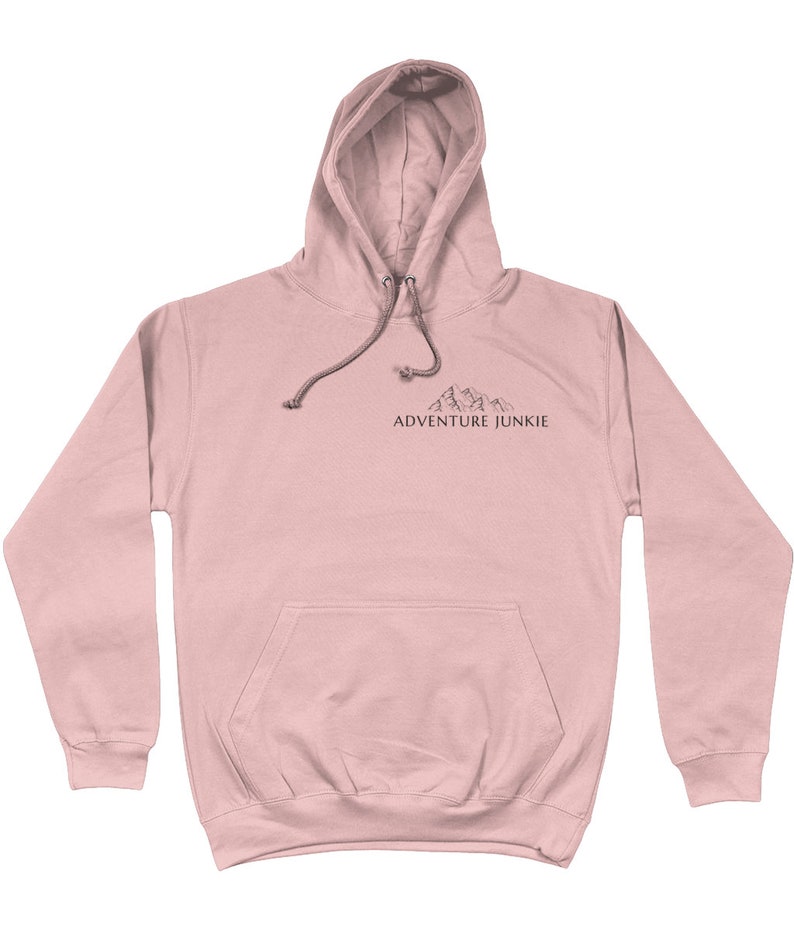 May include: A light pink hoodie with a drawstring hood and a kangaroo pocket. The hoodie features a small mountain graphic and the text "ADVENTURE JUNKIE" printed in black. The hoodie is made of a soft material.