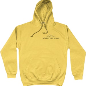 May include: Mustard yellow hoodie with a drawstring hood and a front pocket. The hoodie features a small mountain graphic and the text "ADVENTURE JUNKIE" printed in black.